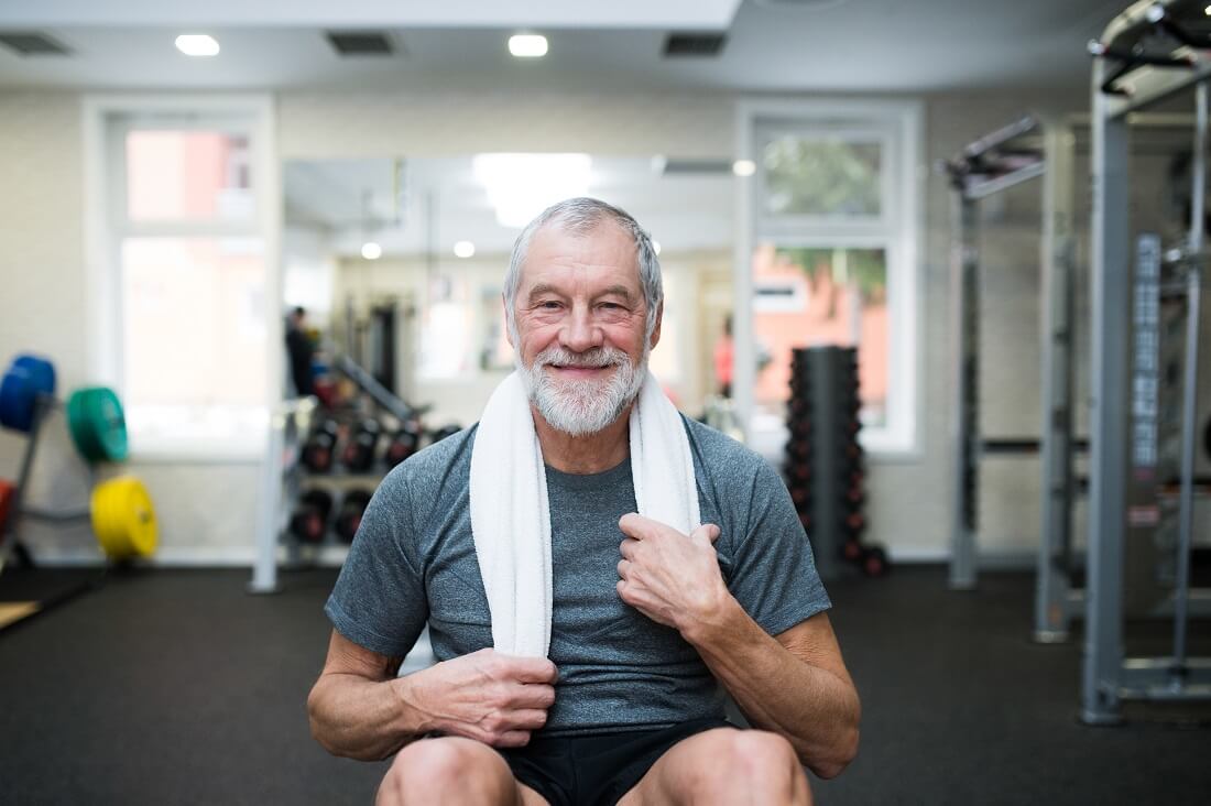 fit senior man in sports clothing in gym resting after working out towel around his neck